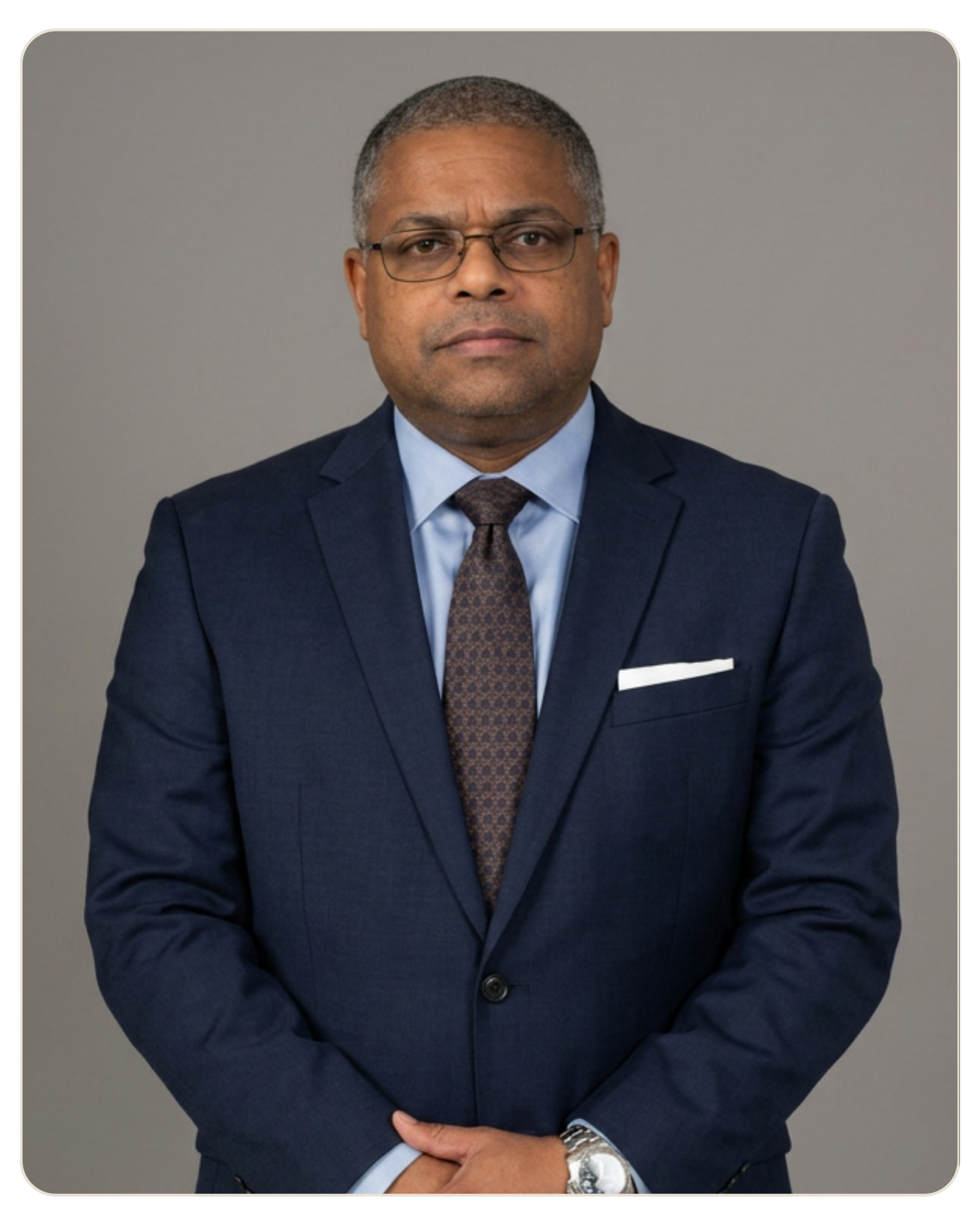 Flyland - Trost - Anthony Bundy, LPC Portrait of a middle-aged man in a navy suit, light blue shirt, brown tie, and glasses against a gray backdrop.