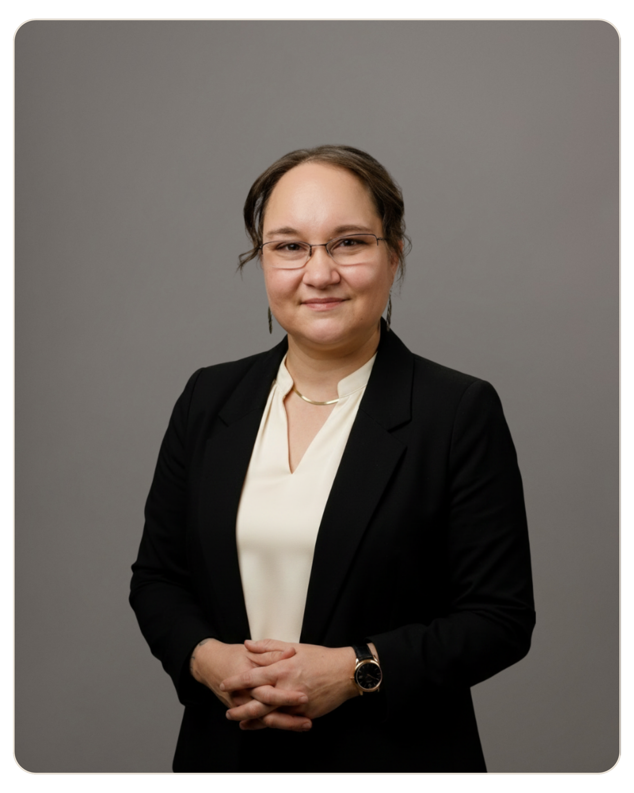 Teri Wilder Professional head-and-shoulders portrait of a woman in a black blazer and light blouse, wearing glasses, with hands folded in front against a gray backdrop.